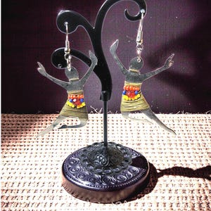 Può includere: Orecchini argentati a forma di ballerina con gonne di perline colorate e braccia tese. Gli orecchini sono esposti su un supporto in metallo nero con una base decorativa, su uno sfondo scuro.