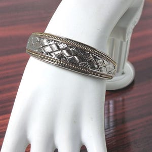 Könnte beinhalten: Ein silberfarbenes Metall-Armband mit einem zentralen Flechtmuster. Das Armband ist von dünnen, goldfarbenen Rändern eingefasst, die entlang der oberen und unteren Kanten des Bandes ein perlenartiges oder strukturiertes Detail aufweisen.