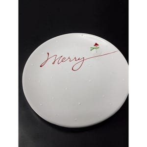 May include: White ceramic plate with the word "Merry" in red script. A small snowman with a red hat and green scarf is on the right side of the plate. Small white dots are scattered across the plate, creating a festive holiday design.