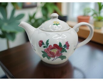 May include: A white ceramic teapot with a lid, featuring a floral design with red and pink flowers, green leaves, and yellow accents. The teapot has a curved handle and spout.