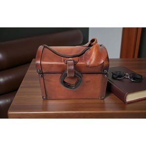 May include: A brown leather chest with a curved top and a handle. The chest has a metal clasp and rivets. Beside it is a closed burgundy book and a pair of sunglasses. The items are on a wooden surface.