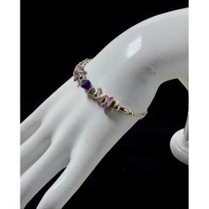 Puede incluir: Una pulsera plateada con una serie de pequeñas piedras irregulares de color púrpura claro y crema, intercaladas con algunas piedras de color púrpura oscuro. La pulsera se muestra en una mano blanca sobre un fondo negro.