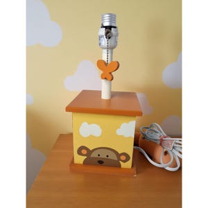 May include: A children's lamp with a yellow and brown wooden base featuring a monkey face and white clouds. The lamp has an orange top and a butterfly-shaped accent. The lamp is on a wooden table.