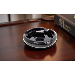 Puede incluir: Un cenicero negro brillante con forma ovalada. El cenicero tiene una superficie lisa y reflectante y un área empotrada para sostener cigarrillos. Un diseño clásico, adecuado para uso doméstico o de oficina.