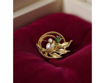 Puede incluir: Broche dorado con una perla, una piedra verde y detalles de hojas. El broche circular tiene un borde texturizado y está sobre un fondo de terciopelo rojo oscuro. Una joya.