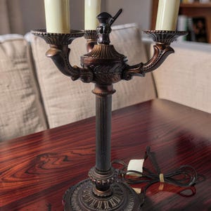May include: A bronze-toned metal candelabra lamp featuring three upright candle-shaped light sockets. The lamp has a fluted central column, a decorative circular base with ornate detailing, and a black electrical cord with a white plug.