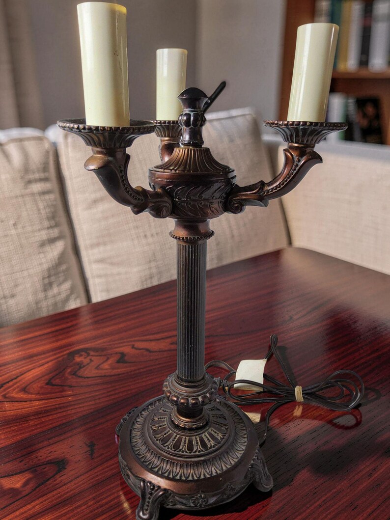 May include: A bronze-toned metal candelabra lamp featuring three upright candle-shaped light sockets. The lamp has a fluted central column, a decorative circular base with ornate detailing, and a black electrical cord with a white plug.