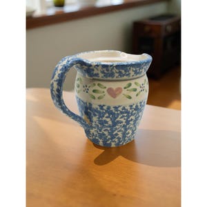 以下が含まれることがあります： 青と白の花柄のセラミックピッチャー。ピッチャーには大きなハンドルと注ぎ口が付いています。緑の葉とピンクのハートの帯が中央を飾っています。ピッチャーは木製のテーブルの上に置かれています。