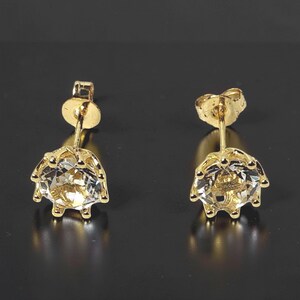 May include: A pair of stud earrings featuring round, clear, faceted gemstones set in gold-toned metal prongs. Each earring has a gold-toned post and a circular butterfly-style backing, showcasing a classic design with a polished metallic finish.