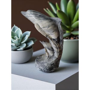 May include: A gray and white marble dolphin sculpture with a textured finish. The dolphin is leaping out of the water, with a second dolphin visible below. The sculpture is displayed on a gray surface, with potted succulents in the background.