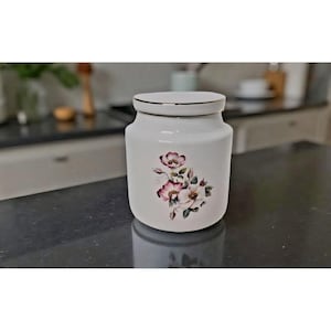 Op de afbeelding: Een witte keramische pot met deksel, versierd met een bloemenpatroon in roze en groen. De pot heeft een gouden rand en staat op een donker aanrecht. De achtergrond is wazig, wat duidt op een keukenomgeving.