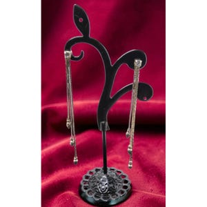 May include: Silver-toned dangle earrings with delicate chains and small, round accents are displayed on a black, decorative earring stand. The stand has a floral-patterned base and a curved, abstract design.