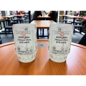 May include: Two clear glass beer mugs with a textured design. Each mug has a printed label with the text "1000 Jahre Mainzer Dom 975-1975" and a black and white illustration of a cathedral. The mugs are on a wooden table.