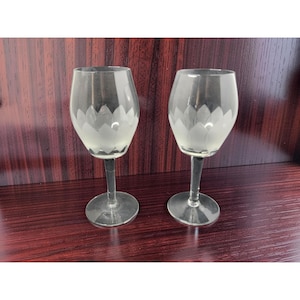 May include: Two clear wine glasses with a frosted floral design on the bowl. The glasses have a long stem and a round base. The background is a dark wood surface.