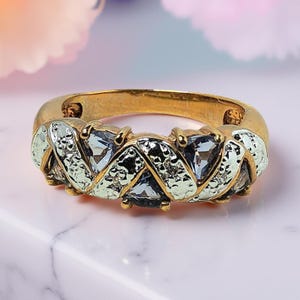 May include: A gold ring featuring light blue gemstones in a textured, interwoven design. The ring's band is gold, and the gemstones are a pale blue. The ring is presented on a white marble surface, showcasing its intricate details.