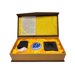 May include: An open, rectangular box with a yellow silk lining contains a black ink stone, a white and blue ceramic ink well, and a black ink block. A paper with Chinese characters is in the box lid.