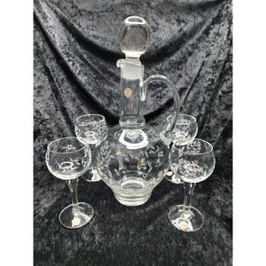 May include: A clear glass decanter with a stopper and a handle, surrounded by five matching wine glasses. The decanter and glasses feature an etched floral design. The set is displayed on a dark, textured surface.