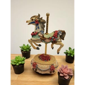 May include: A carousel horse figurine with a cream-colored body, adorned with red and blue floral accents. The base is cream-colored with red roses. Small potted succulents surround the carousel horse on a wooden surface.