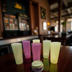 May include: A collection of colorful plastic tumblers in shades of yellow, purple, and green, arranged on a dark wooden table. A stack of coasters in matching colors sits in front of the glasses. The background shows a bar setting.