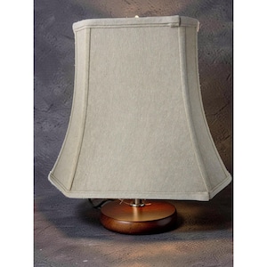 May include: A small table lamp featuring a beige fabric lampshade and a dark brown wooden base. The lampshade has a flared shape and is rectangular at the top. The base is round and has a polished finish. The lamp is switched on, and the light is visible.
