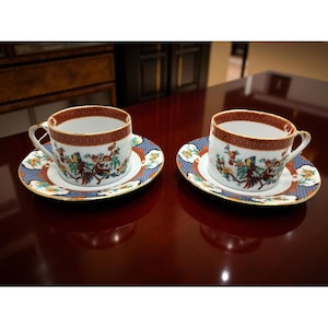 May include: Two matching teacups and saucers. The cups have a white interior and a decorative band of red and gold around the rim. The saucers have a blue and white floral pattern with a gold trim. The cups and saucers are placed on a dark wood surface.