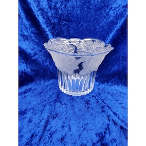 Può includere: Un portacandele in vetro trasparente con un design floreale smerigliato intorno alla parte superiore. La base ha delle scanalature verticali. Il portacandele è posto su uno sfondo blu.