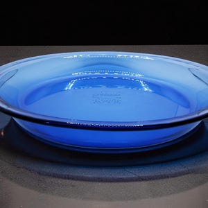 May include: A round, transparent blue glass pie dish with a wide, flat rim. The center of the dish features the embossed text "pyrex" followed by "MADE IN U.S.A." and "FOR OVEN AND MICROWAVE" in small, clear, centered lettering on the bottom surface.
