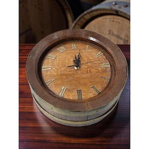 Könnte beinhalten: Eine runde Holzuhr mit hellbraunem Zifferblatt und dunkelbraunem Rahmen. Die Uhr hat römische Ziffern und schwarze Zeiger. Die Uhr ist aus Holz gefertigt und hat ein rustikales Aussehen, mit Metallbändern an den Seiten.