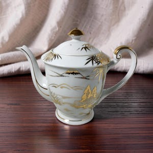 May include: A white porcelain teapot featuring gold-coloured illustrations of Mount Fuji, bamboo stalks, and landscape scenery. The teapot includes a matching lid with a gold-coloured finial, a curved spout, and a gold-accented handle on the side.