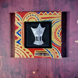 May include: A silver-toned metal incense burner pin featuring an intricate, pointed top design with engraved details and vertical bars, set against a black background within a square frame painted with red, blue, and gold geometric patterns.