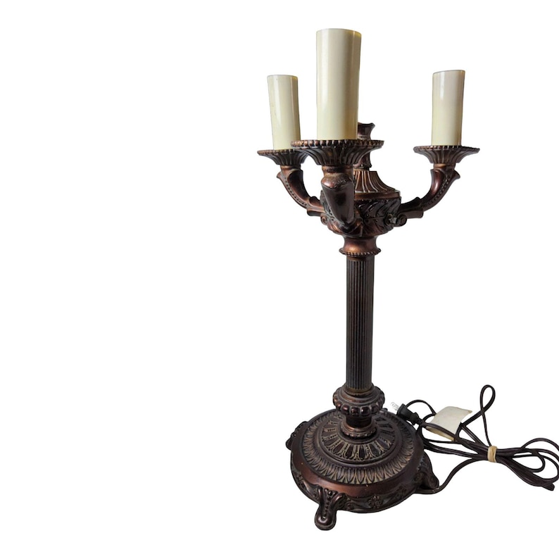 May include: A three-light candelabra lamp with a dark bronze-toned finish. The lamp features a fluted central column, an ornate circular base, and three cream-colored cylindrical light covers. A black electrical cord extends from the base.