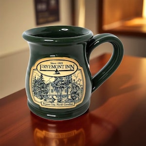 Peut inclure: Mug en céramique vert foncé avec une anse incurvée. La tasse présente une étiquette dorée avec le texte "FRYEMONT INN Since 1923" et une illustration d'un bâtiment et d'arbres. Le texte "Bryson City, North Carolina" est également sur l'étiquette.