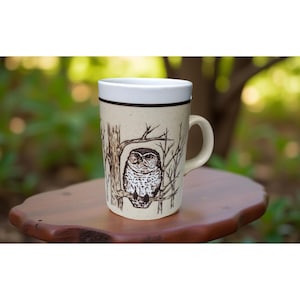 May include: A beige ceramic mug featuring a detailed illustration of an owl perched on a tree branch. The mug has a white rim and a brown band near the top. The background shows a blurred green foliage.