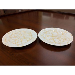 Può includere: Due piatti da portata ovali bianchi con un disegno a scarabocchi dorati. I piatti sono posizionati su una superficie di legno marrone scuro. I piatti sono vuoti e pronti per servire il cibo.