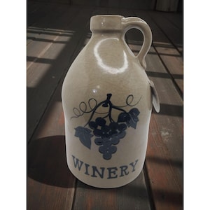 May include: A beige ceramic jug with a handle, featuring a blue grape design and the word "WINERY". The jug has a crackled finish and a tag attached. It is a decorative item.