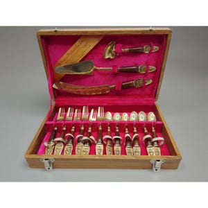 May include: A vintage wooden cutlery set with a pink velvet interior. The set includes forks, spoons, knives, and serving utensils with ornate handles and silver-colored blades. The case is closed with two metal clasps.