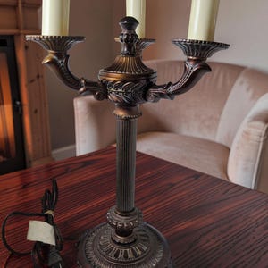 May include: A three-light candelabra lamp with a dark bronze finish, featuring a fluted central column, ornate base, and three cream-colored candle-style light covers. The lamp includes a black electrical cord with a standard two-prong plug.