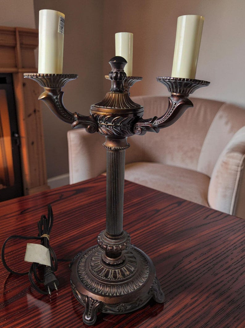 May include: A three-light candelabra lamp with a dark bronze finish, featuring a fluted central column, ornate base, and three cream-colored candle-style light covers. The lamp includes a black electrical cord with a standard two-prong plug.
