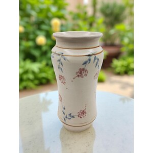 May include: A cream ceramic vase with a gently curved shape, featuring a flared top and base. It is decorated with floral patterns in blue and pink. The design includes small clusters of pink flowers and blue leaves, adding a touch of elegance.