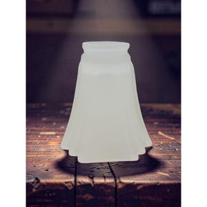 May include: A frosted glass lampshade with a flared, skirt-like design. The lampshade is opaque and sits on a dark wooden surface. The lampshade has a classic, vintage aesthetic.