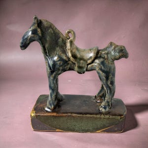 May include: A ceramic horse figurine featuring a mottled blue, gray, and brown glaze. The horse stands on a rectangular base and includes a saddle detail on its back. The finish is glossy with variations in color throughout the surface of the piece.