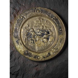May include: A decorative brass plate with a detailed relief scene of people gathered around a table. The plate has an ornate, floral border and a dark, textured background. The relief depicts figures in period clothing.