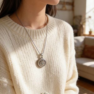 May include: A silver pendant necklace featuring a circular medallion with a central flower design and beaded border, worn over a cream-colored knit sweater. Two small circular pins are attached to the sweater, one with a floral motif and one with the text Get Good.