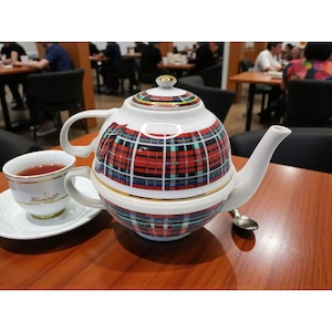 May include: A white ceramic teapot and teacup set with a red, green, and blue plaid pattern. The teapot has a gold rim and a matching lid. A teacup with tea and a saucer are next to the teapot. A small spoon rests on the table.