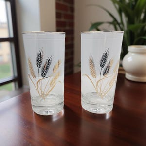 May include: Two clear, tall drinking glasses with a wheat stalk design. The wheat stalks are in black, gold, and brown, with gold rims. The glasses are on a dark brown wooden surface.