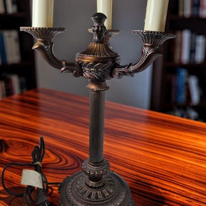 May include: A bronze-toned metal candelabra lamp featuring a fluted central column, a decorative circular base, and three arms holding off-white cylindrical candle-style bulbs. A black electrical cord with a plug is attached to the base.
