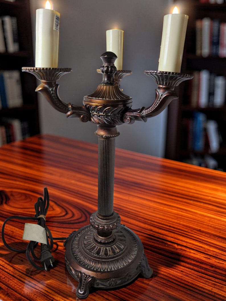 May include: A bronze-toned metal candelabra lamp featuring a fluted central column, a decorative circular base, and three arms holding off-white cylindrical candle-style bulbs. A black electrical cord with a plug is attached to the base.
