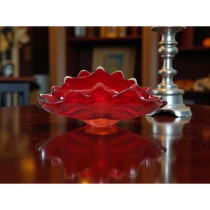 May include: A vibrant red glass bowl with a ruffled edge and a white trim. The bowl is round and sits on a small base. The bowl is reflecting on a dark brown surface. A silver candlestick holder is in the background.