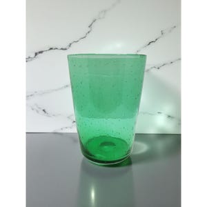 May include: A tall, green glass with a slightly textured surface. The glass has a cylindrical shape and is filled with small, evenly spaced bubbles. The glass is set against a white and grey marble-patterned background.