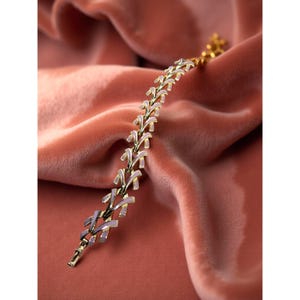 May include: A gold-toned bracelet with a delicate, repeating leaf design. The leaves are accented with a pale lavender enamel. The bracelet is displayed on a soft, rose-colored fabric, creating a luxurious feel.